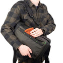 A person with the Think Tank Backlight Charcoal Colored Backpack resting on his waist as he pulls out a hat out of a pocket of the bag.